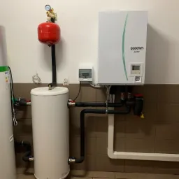 Biały zbiornik buforowy z czerwonym naczyniem przeponowym, podłączony do pompy ciepła Ecodan, widoczne rury z izolacją termiczną i osprzęt, na tle brązowych płytek.