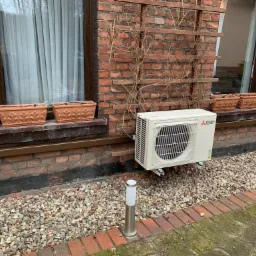 Zewnętrzna jednostka klimatyzacji Mitsubishi Electric zamontowana na ceglanej ścianie domu z dwoma oknami, na tle żwirowego podłoża i niskiej lampy ogrodowej.