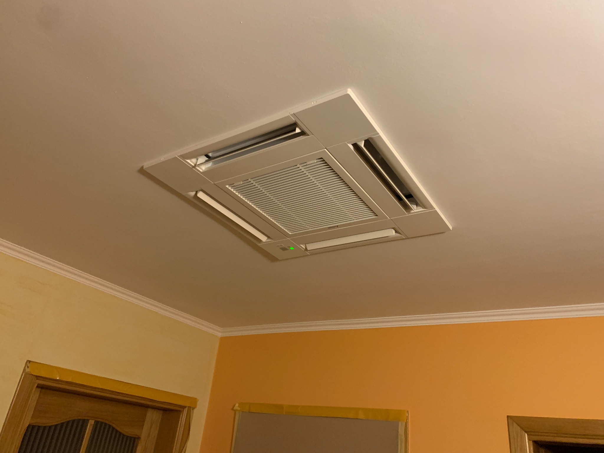 Wbudowana w sufit kaseta klimatyzacyjna z widoczną kratką wentylacyjną i zieloną diodą sygnalizacyjną, zamontowana w pomieszczeniu z żółto-pomarańczowymi ścianami i ozdobnymi listwami przysufitowymi.