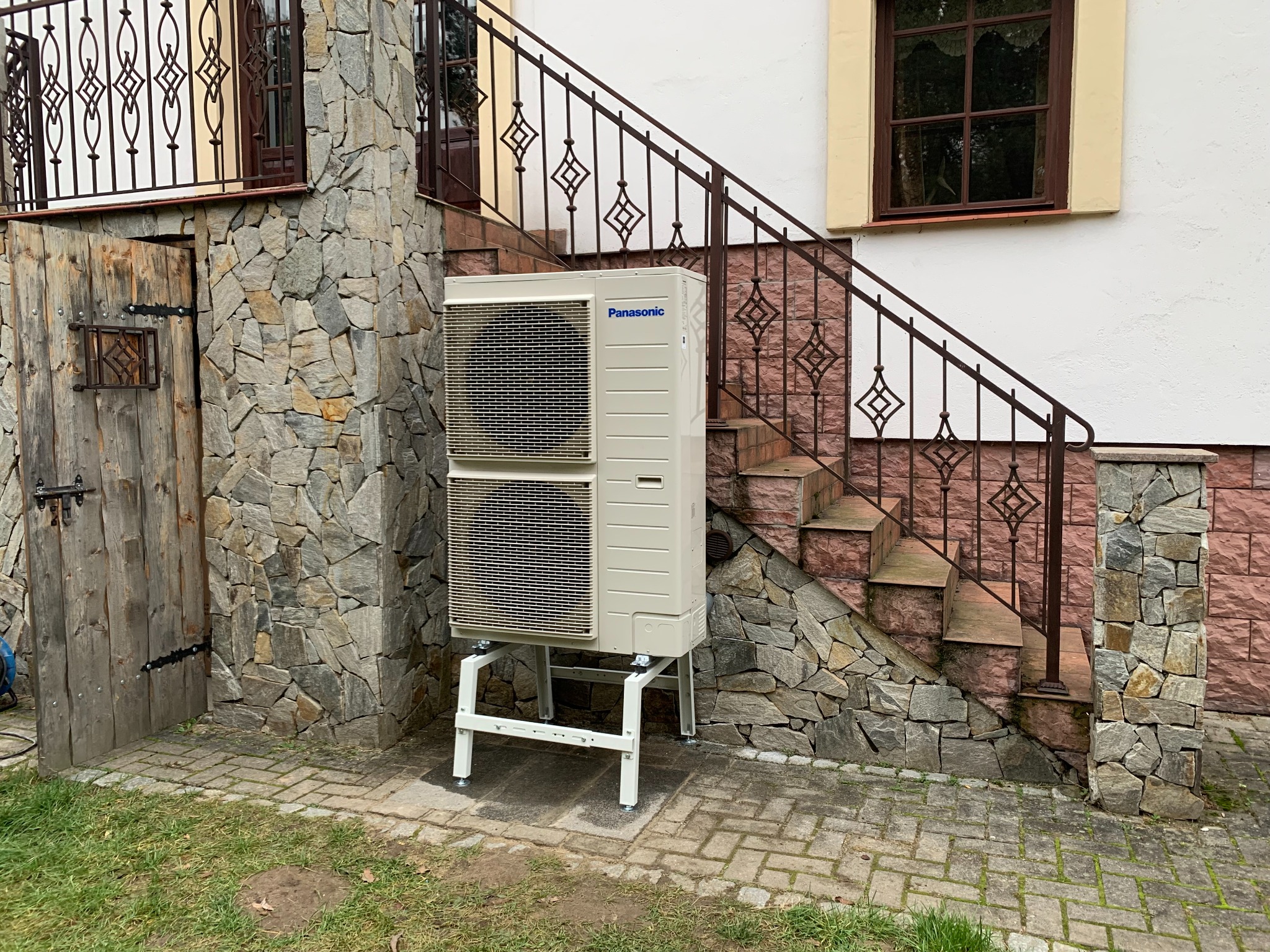 Zewnętrzna jednostka klimatyzacji Panasonic na białym stelażu, ustawiona na kostce brukowej obok kamiennej ściany budynku z drewnianymi drzwiami i schodami z metalową balustradą.