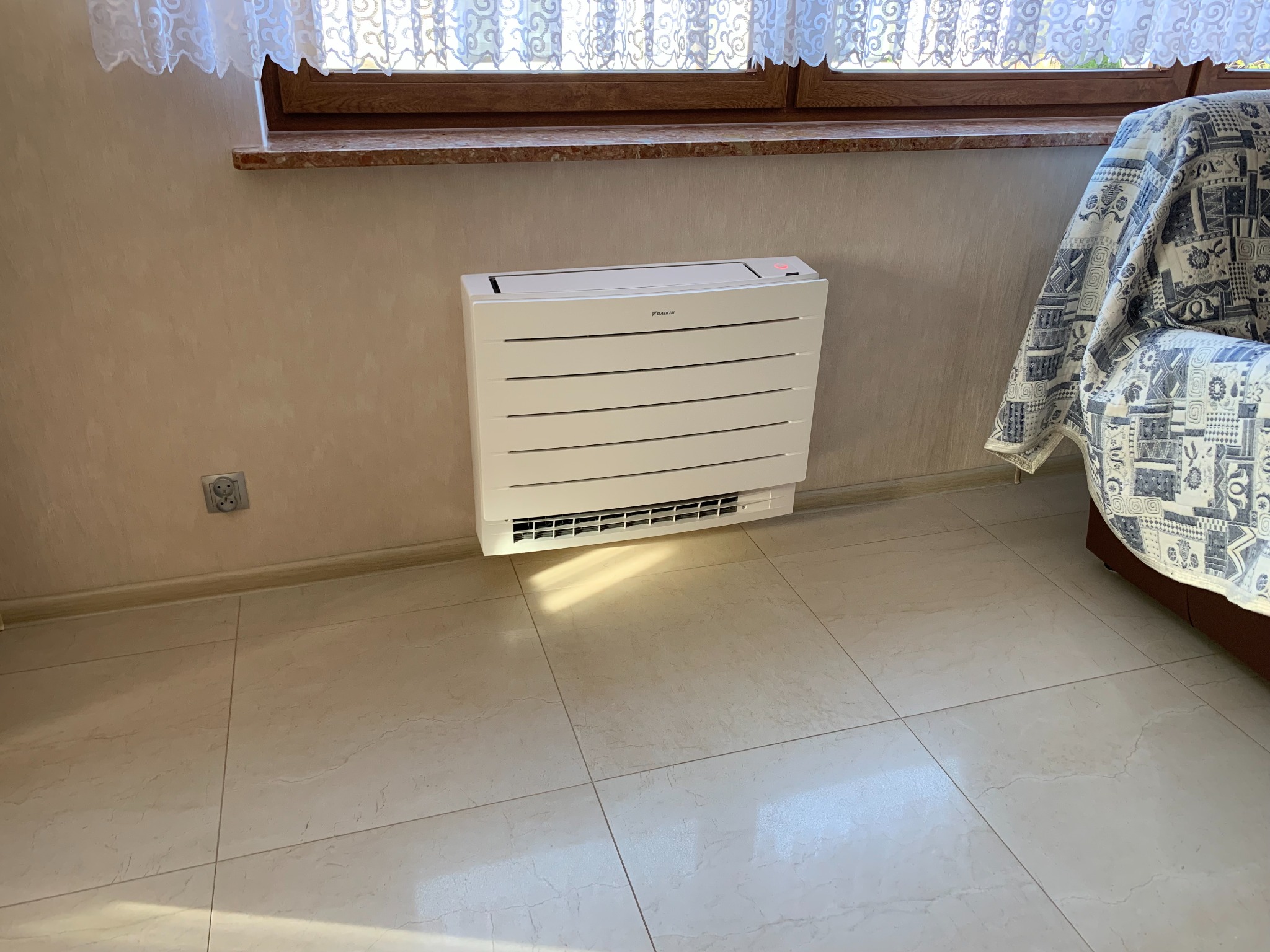 Biały klimatyzator podłogowy Daikin zamontowany przy ścianie w jasnym pomieszczeniu z marmurową podłogą i oknem z koronkową firanką.