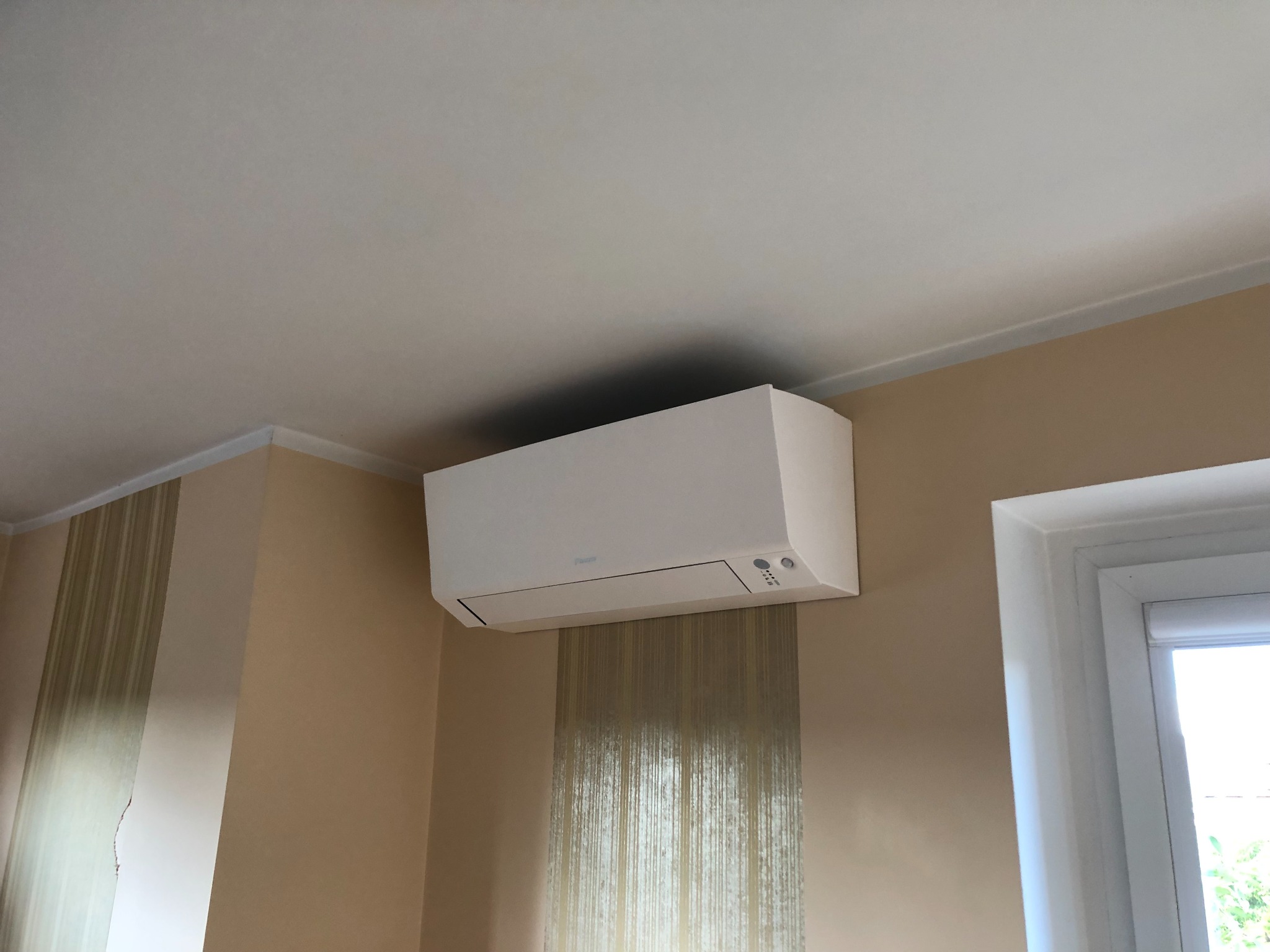 Biała jednostka klimatyzacji Daikin zamontowana na ścianie z beżową tapetą w mieszkaniu, widoczny fragment okna z białymi ramami.