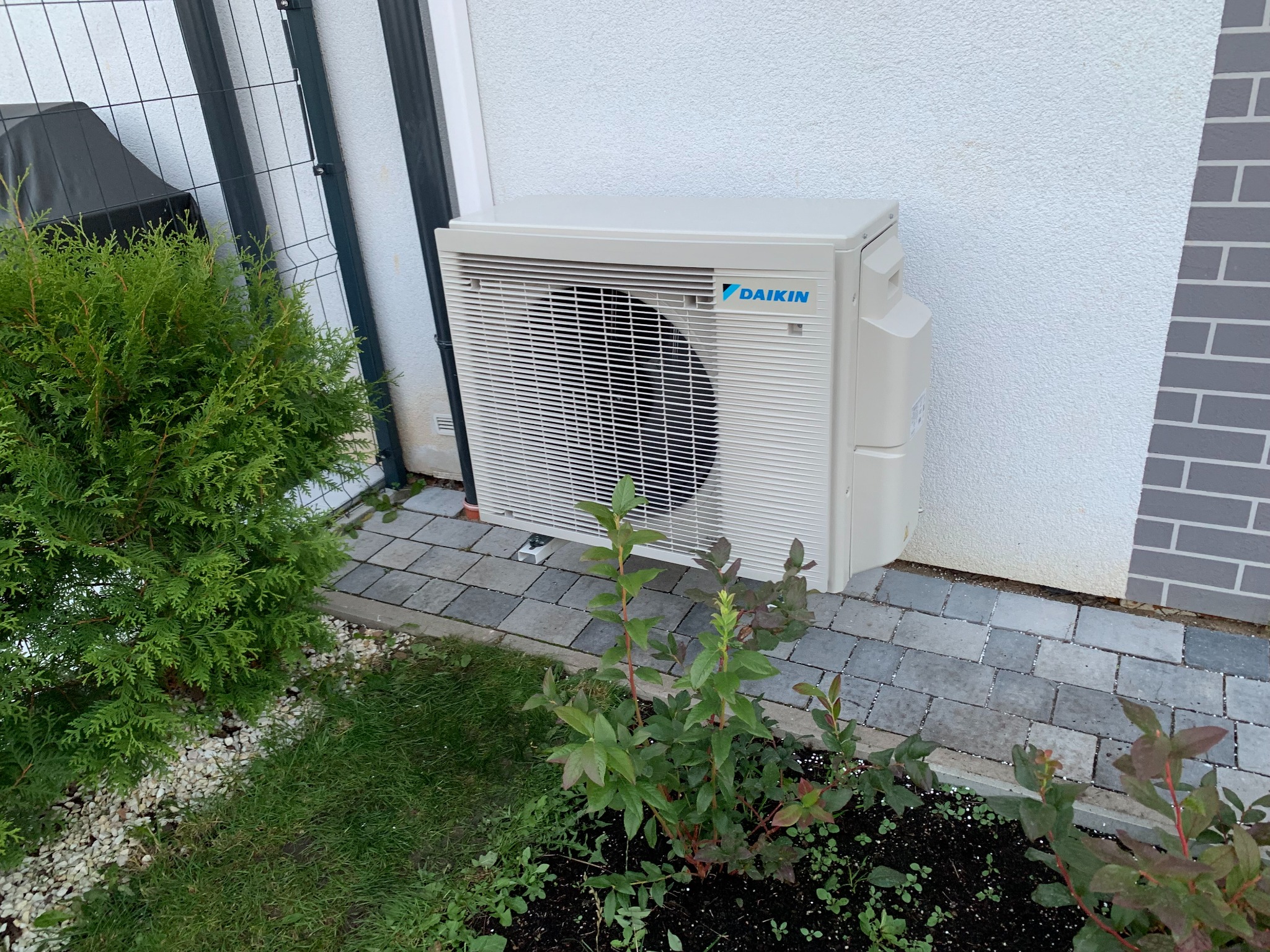 Zewnętrzna jednostka klimatyzacji Daikin zamontowana przy ścianie domu, obok zadbanego ogrodu z tujami i kostką brukową.