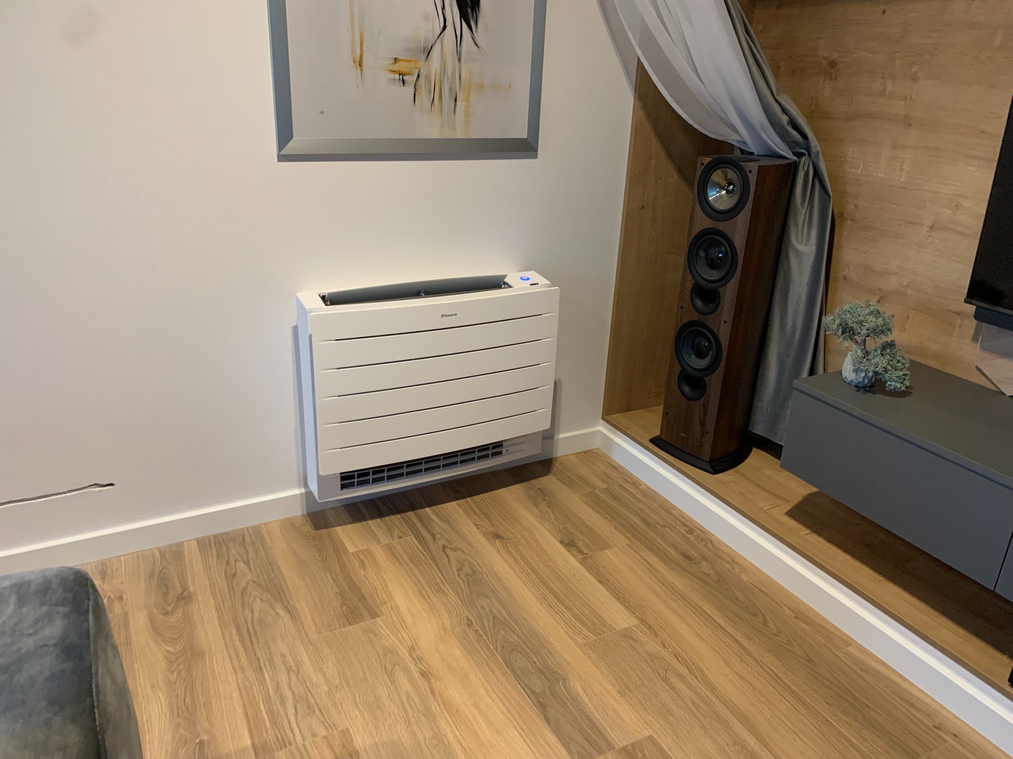 Biały klimatyzator podłogowy Daikin w nowoczesnym salonie z drewnianą podłogą, obok kolumna głośnikowa i szara komoda z dekoracyjnym bonsai, abstrakcyjny obraz na ścianie.
