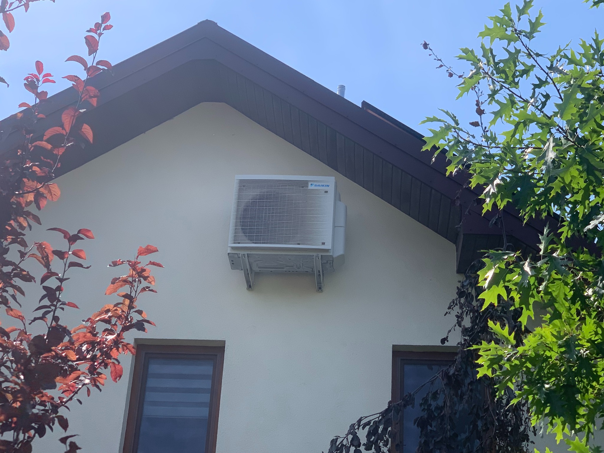 Biała jednostka zewnętrzna klimatyzacji Daikin zamontowana na jasnej ścianie domu jednorodzinnego, widoczna z dołu, z elementami zieleni i czerwonych liści w tle.
