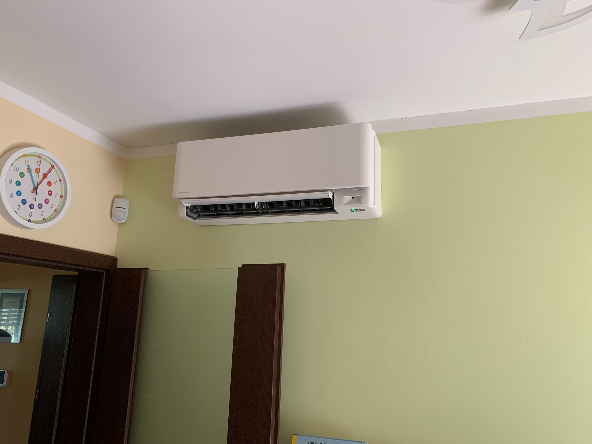 Biała jednostka klimatyzacyjna marki Daikin zamontowana na ścianie w pokoju o pastelowych ścianach, widoczny zegar z kolorowymi cyframi i fragment ciemnobrązowej framugi drzwi.