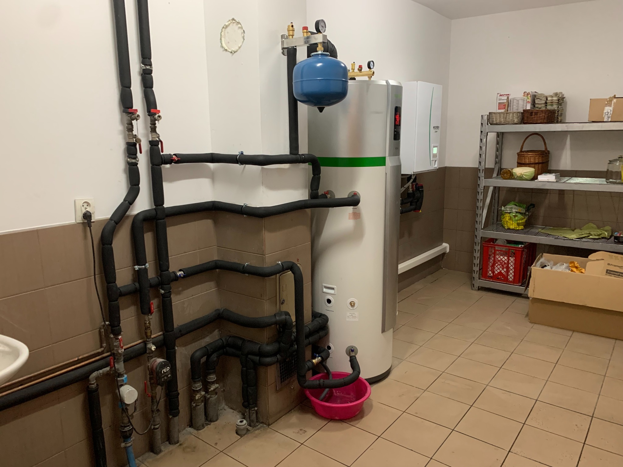 Instalacja rekuperacji z zbiornikiem akumulacyjnym, widoczne rury w izolacji, manometry, zawory oraz półka z zapasami w tle.