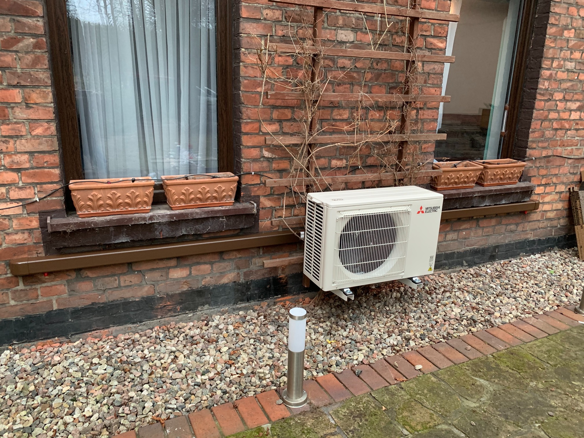 Zewnętrzna jednostka klimatyzacji Mitsubishi Electric zamontowana na ceglanej ścianie domu z dwoma oknami, na tle żwirowego podłoża i niskiej lampy ogrodowej.