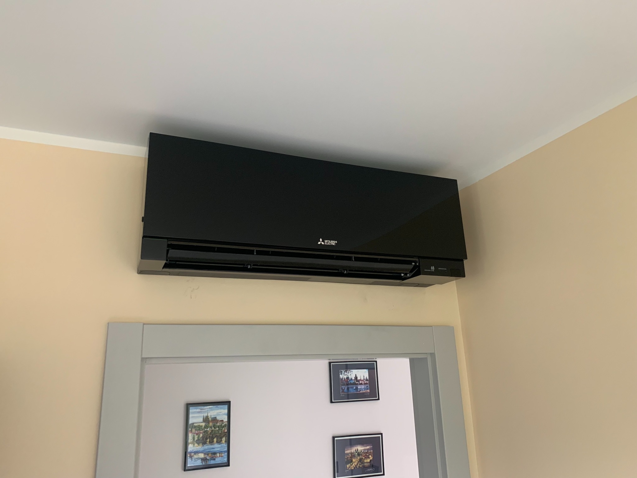 Czarny klimatyzator Mitsubishi Electric zamontowany na ścianie nad szarym obramowaniem drzwi, z trzema małymi obrazkami na ścianie w tle.