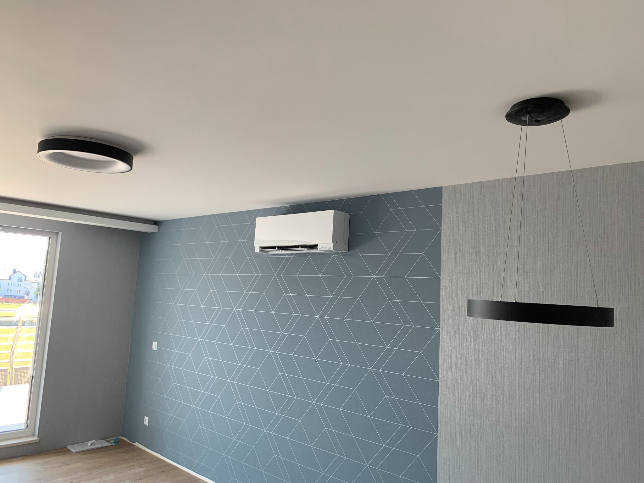 Nowoczesne wnętrze z zamontowanym klimatyzatorem ściennym, okrągłą lampą sufitową i wiszącą lampą pierścieniową na tle ściany z geometrycznym wzorem i teksturowanej ściany.