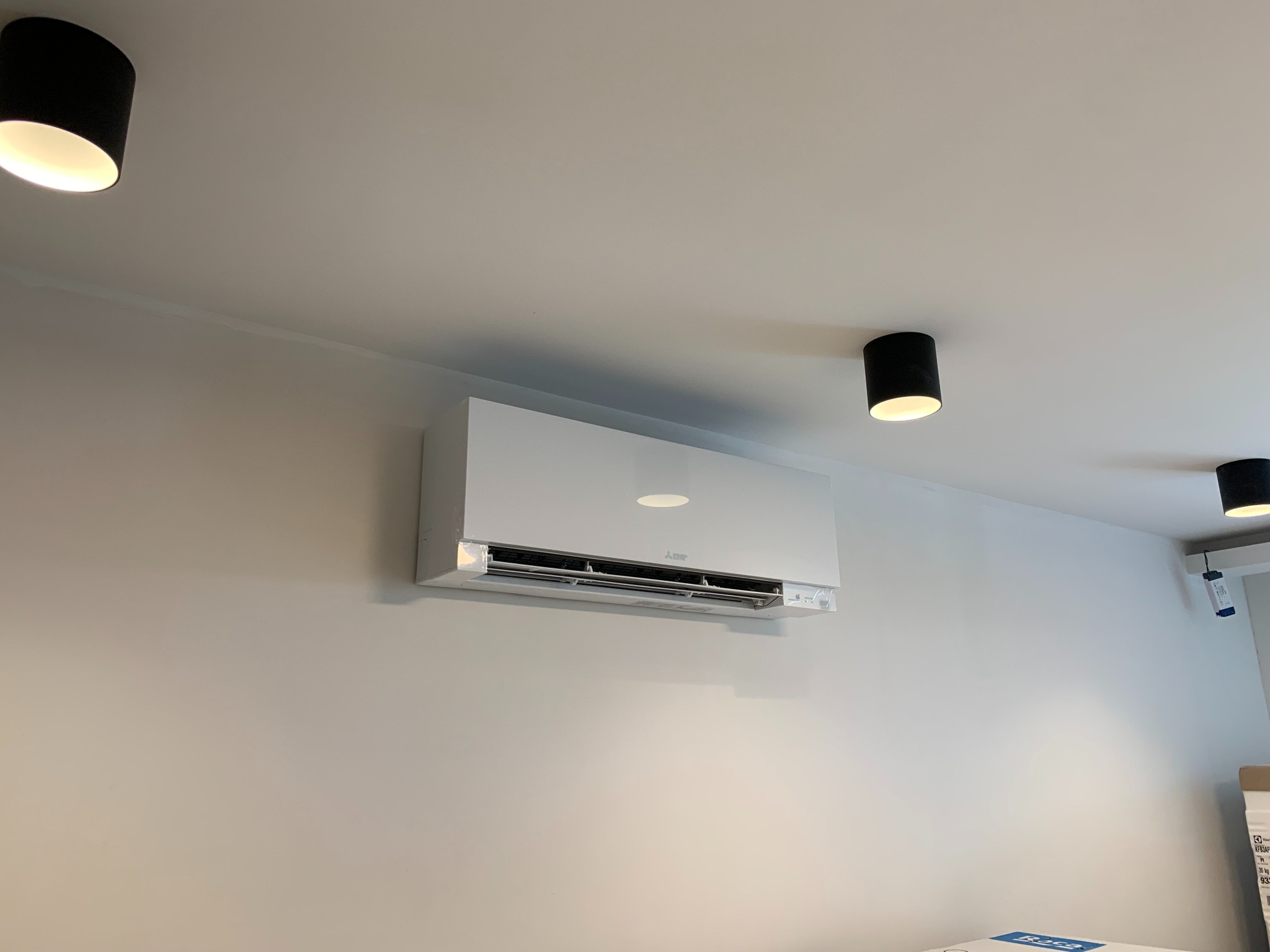 Biały klimatyzator Mitsubishi Electric zamontowany na jasnej ścianie w nowoczesnym wnętrzu, widoczne trzy czarne, okrągłe lampy sufitowe.