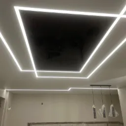 Nowoczesny sufit z podświetleniem LED w kształcie geometrycznych figur, czarna, błyszcząca wstawka w centralnej części, wiszące lampy w folii ochronnej, widoczny fragment osoby w lewym dolnym rogu.