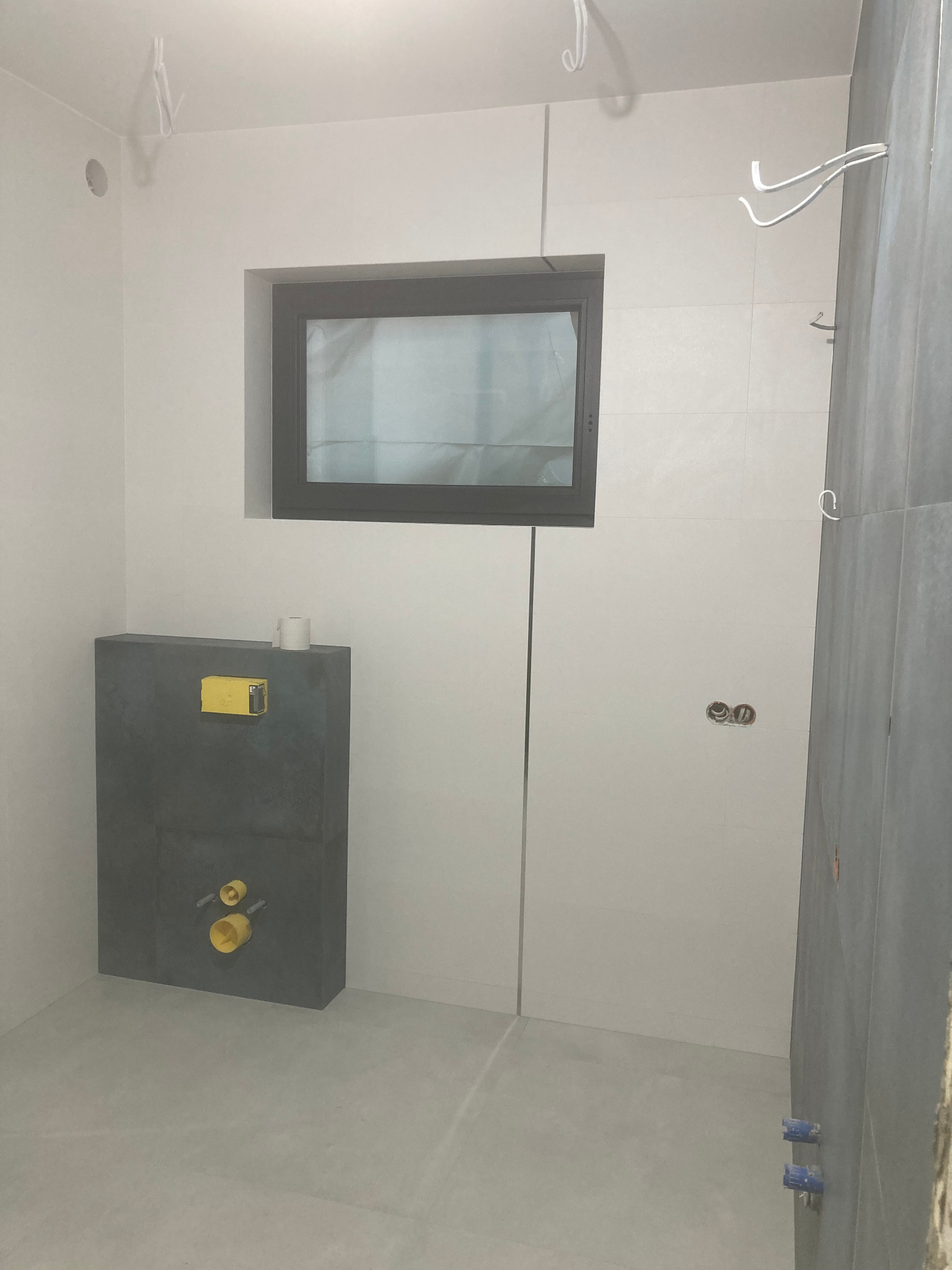 Nowoczesna łazienka w trakcie wykańczania: szare płytki na ścianie i podłodze, podtynkowa instalacja WC z szarą obudową, okno z czarną ramą, wystające kable elektryczne i rury hydrauliczne.