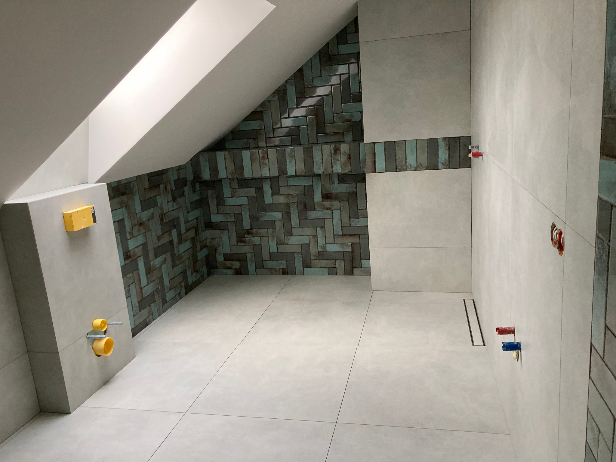 Ścianka kolankowa w łazience na poddaszu z ułożoną częściowo mozaiką w odcieniach szarości i turkusu, widoczne przyłącza wodne i odpływ liniowy w podłodze.