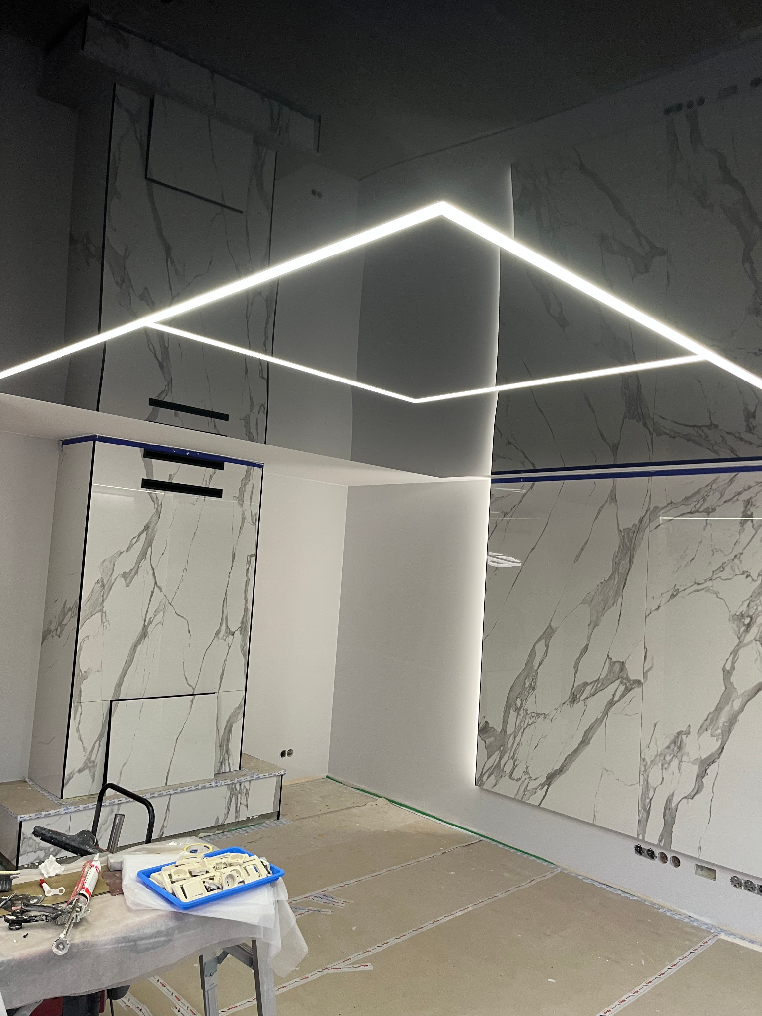 Nowoczesne wnętrze w trakcie remontu, z błyszczącym, czarnym sufitem napinanym podświetlonym geometrycznymi liniami LED, ściany wyłożone płytami imitującymi marmur, widoczne narzędzia i akcesoria...