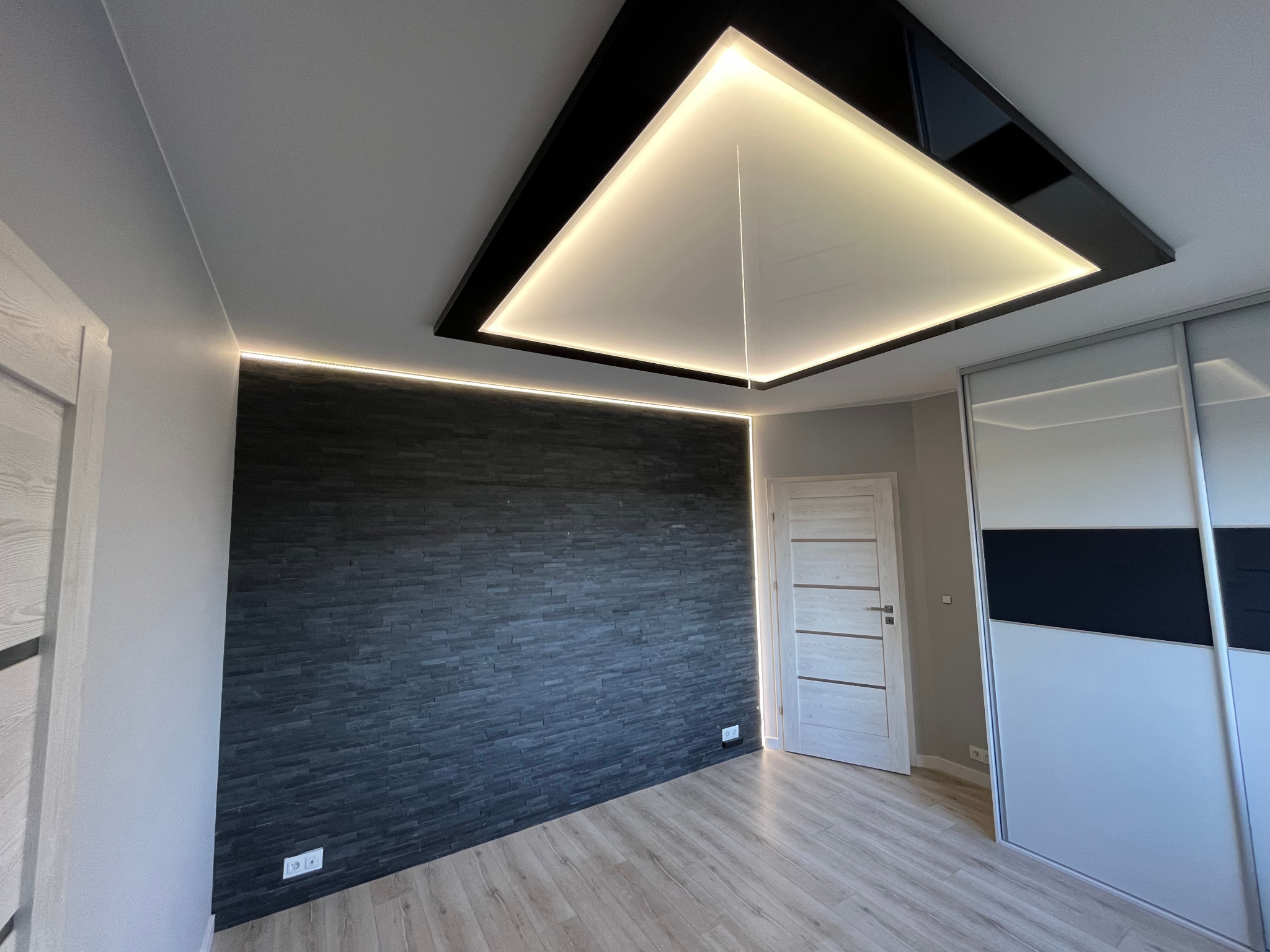 Nowoczesne wnętrze pokoju z czarną ścianą z kamienia dekoracyjnego, podświetlanym sufitem podwieszanym z czarną obudową i oświetleniem LED, oraz panele podłogowe imitujące drewno.