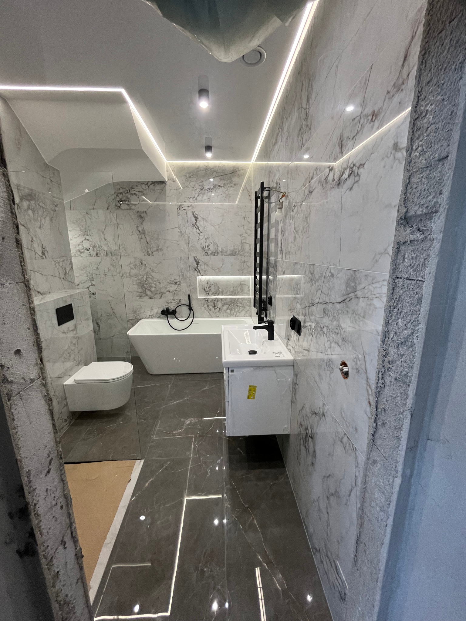 Wnętrze łazienki w trakcie remontu, z marmurowymi płytkami na ścianach, nowoczesną białą wanną, umywalką i toaletą w zabudowie, widoczne niedokończone elementy wykończeniowe.