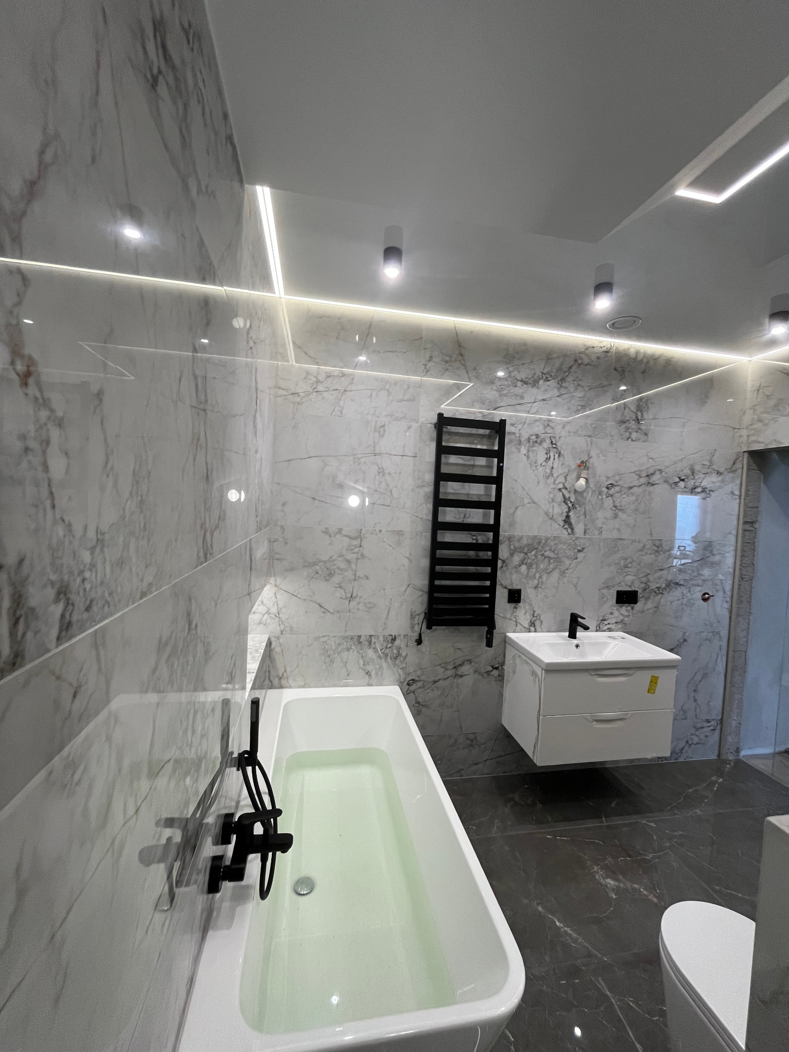 Nowoczesna łazienka z marmurowymi płytkami, podświetleniem LED, wiszącą szafką z umywalką i czarną armaturą, grzejnikiem drabinkowym oraz wanną wypełnioną wodą.