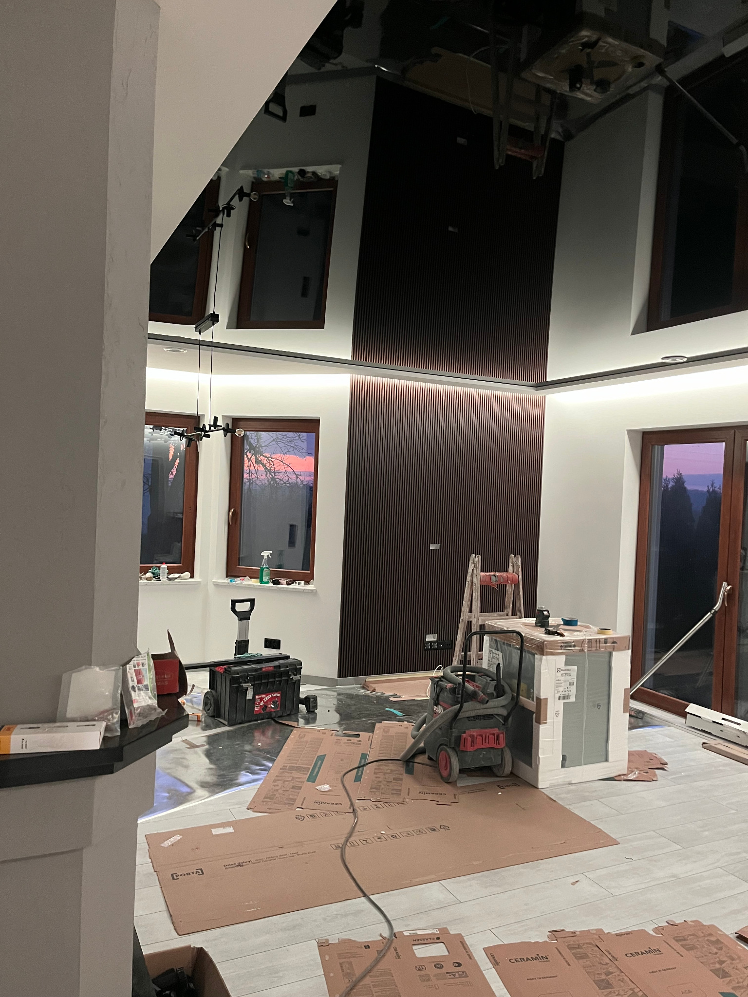 Wnętrze w trakcie wykończenia: ściana z paneli dekoracyjnych, częściowo zabezpieczona podłoga, narzędzia, kartony po panelach podłogowych, drabina oparta o ścianę, widok na zachód słońca przez okno.