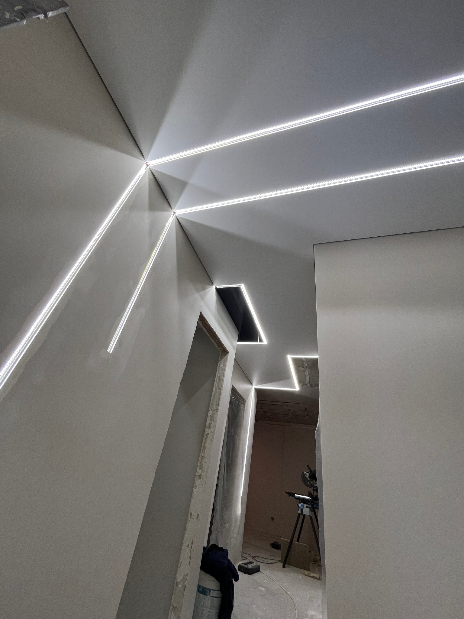 Nowoczesny sufit z liniowym oświetleniem LED tworzącym geometryczne wzory, widoczne niedokończone wykończenia ścian i fragment narzędzi w tle.