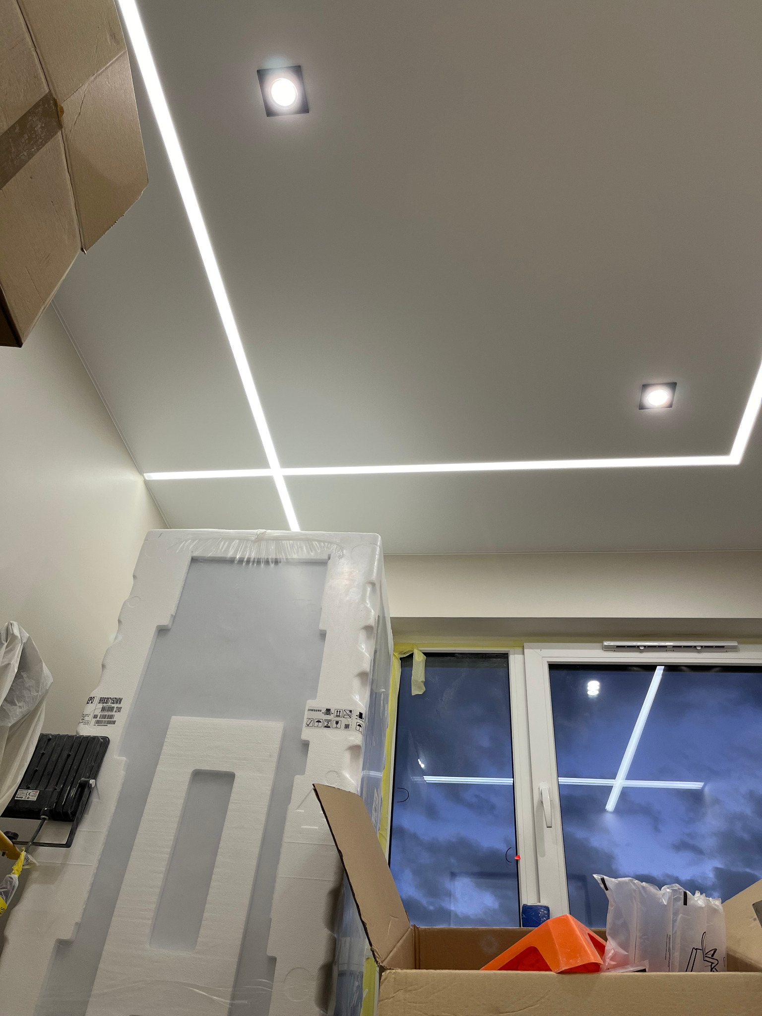 Biały sufit z liniowym oświetleniem LED tworzącym geometryczny wzór, widoczne kwadratowe lampy punktowe, okno z widokiem na pochmurne niebo oraz materiały budowlane w opakowaniach, wskazujące...