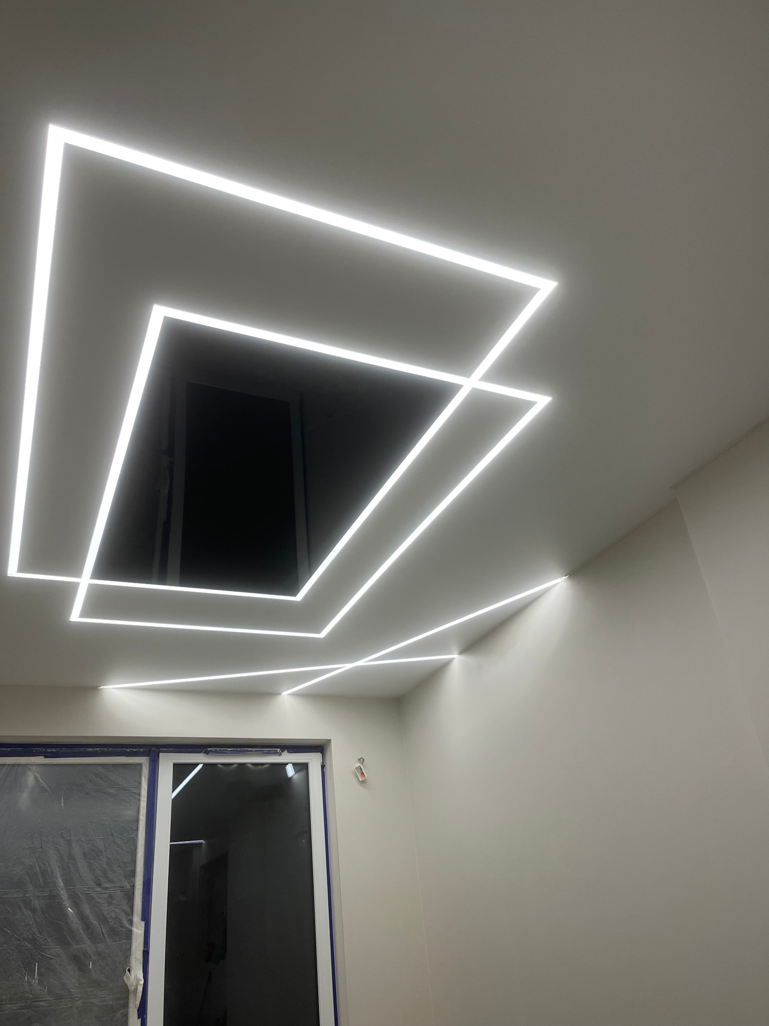 Nowoczesny sufit z geometrycznym oświetleniem LED w kształcie kwadratów, widoczne okno zabezpieczone folią malarską, ściany w jasnym odcieniu szarości.