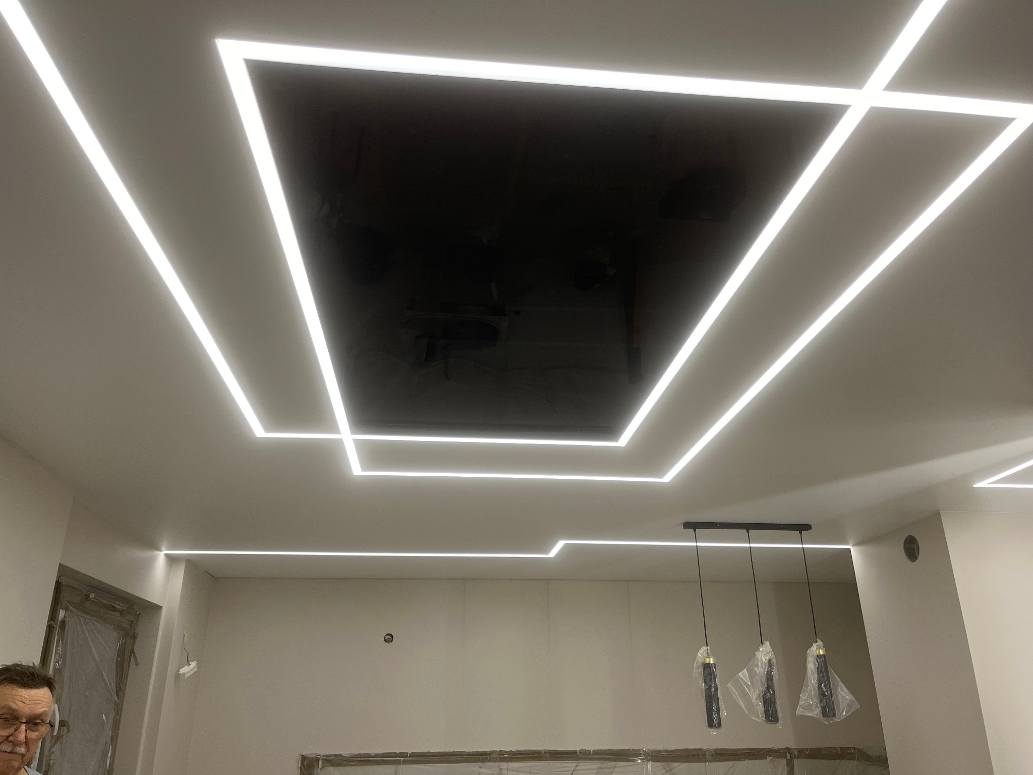 Nowoczesny sufit z podświetleniem LED w kształcie geometrycznych figur, czarna, błyszcząca wstawka w centralnej części, wiszące lampy w folii ochronnej, widoczny fragment osoby w lewym dolnym rogu.