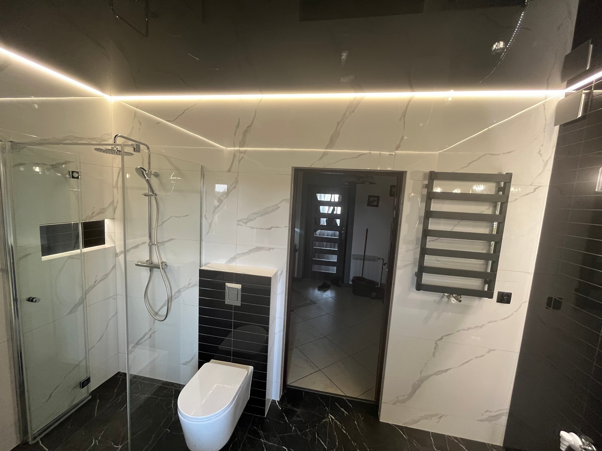 Nowoczesna łazienka z czarno-białą marmurową glazurą, podświetlanym sufitem napinanym, kabiną prysznicową typu walk-in i czarnym grzejnikiem drabinkowym.