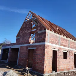 Surowy stan domu jednorodzinnego z czerwonej cegły, widoczna konstrukcja dachu z drewnianych belek, niezadaszony garaż z boku, błękitne niebo w tle.