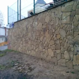 Kamienny mur z nieregularnych, brązowo-beżowych płyt, fragment muru z czerwonej cegły z prawej strony. W tle siatka ogrodzeniowa i drzewo bez liści.