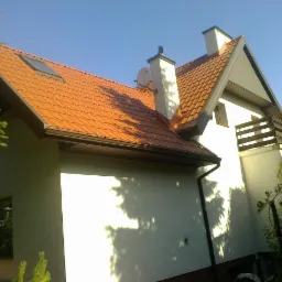 Dom jednorodzinny z pomarańczową dachówką, widoczne okno dachowe, kominy i rynny. Ujęcie z dołu, błękitne niebo w tle, częściowo ocieniony elewacja.