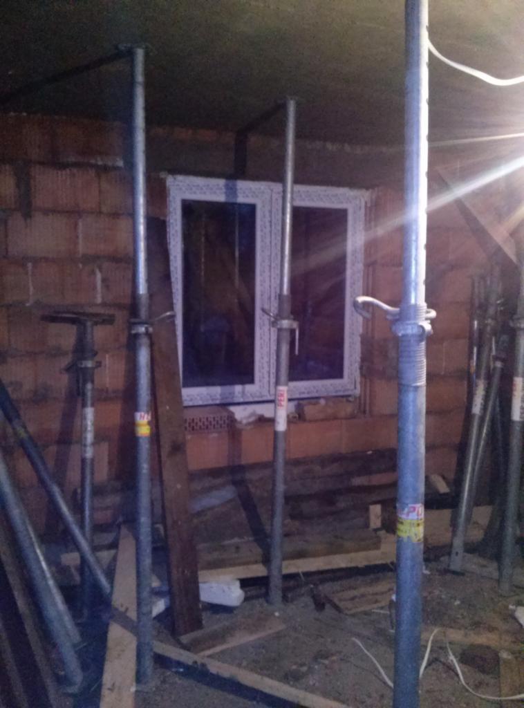 Montaż okna w surowym murze, widoczne podpory budowlane i elementy drewniane. Oświetlenie boczne podkreśla fakturę cegły.