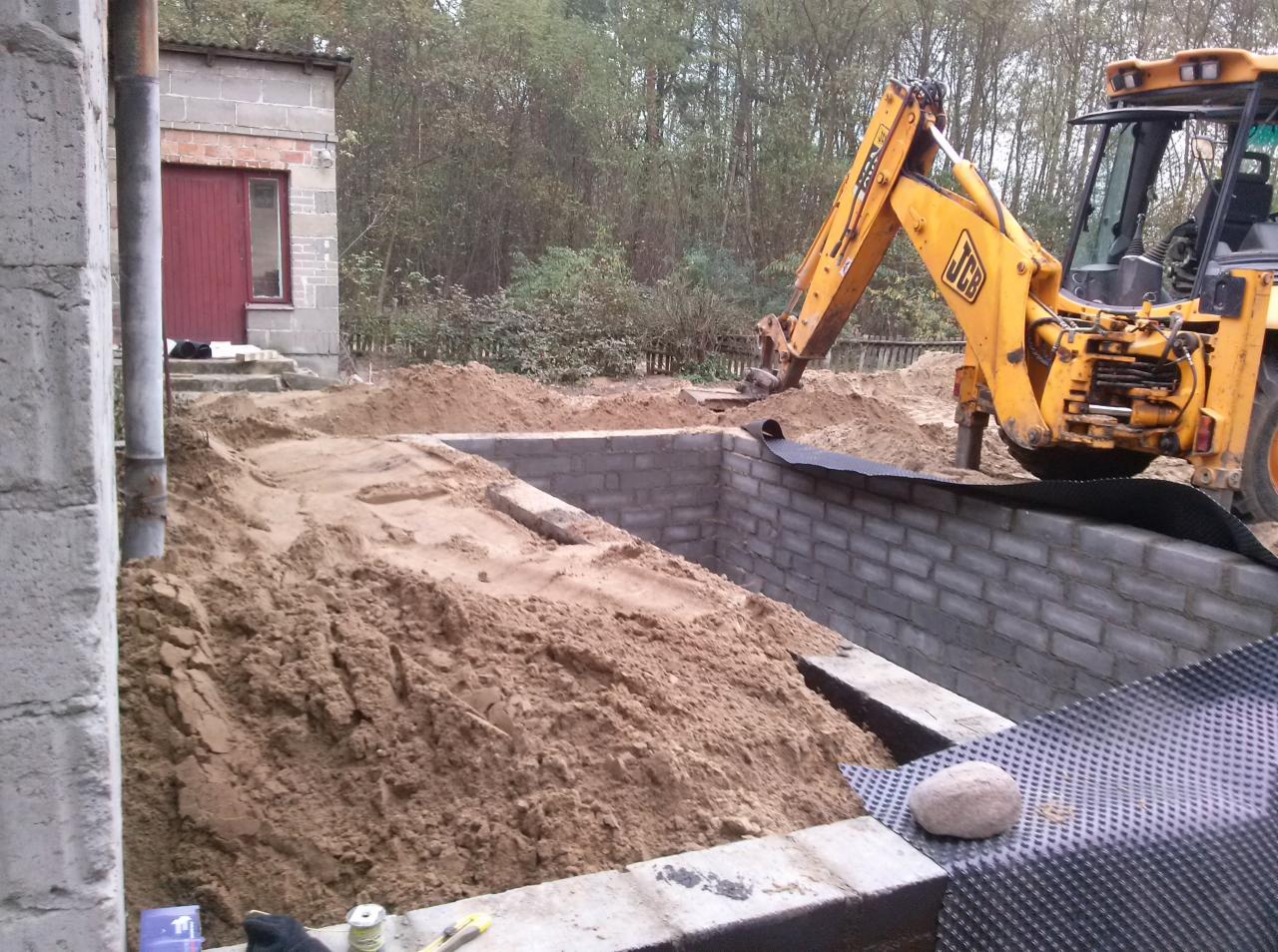 Fundament z bloczków, zabezpieczony folią, zasypywany piaskiem przez koparko-ładowarkę JCB. Widoczny fragment budynku w tle. Prace ziemne, etap budowy.
