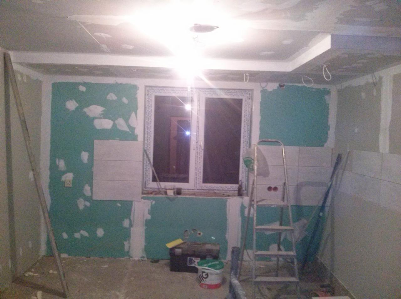 Wnętrze w trakcie remontu: płyta karton-gips na ścianach, częściowo położone płytki, okno PCV. Widoczne narzędzia i drabina. Etap prac wykończeniowych.