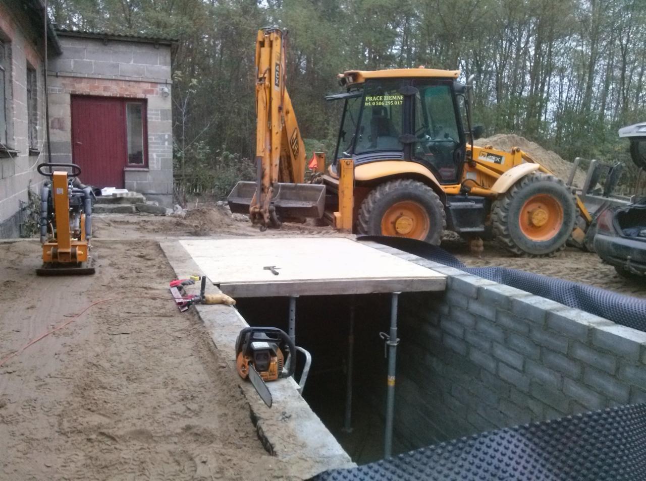 Fundament budynku w trakcie budowy, zabezpieczony folią kubełkową, z widocznym wykopem i sprzętem budowlanym: koparka, zagęszczarka, piła spalinowa. Prace ziemne.