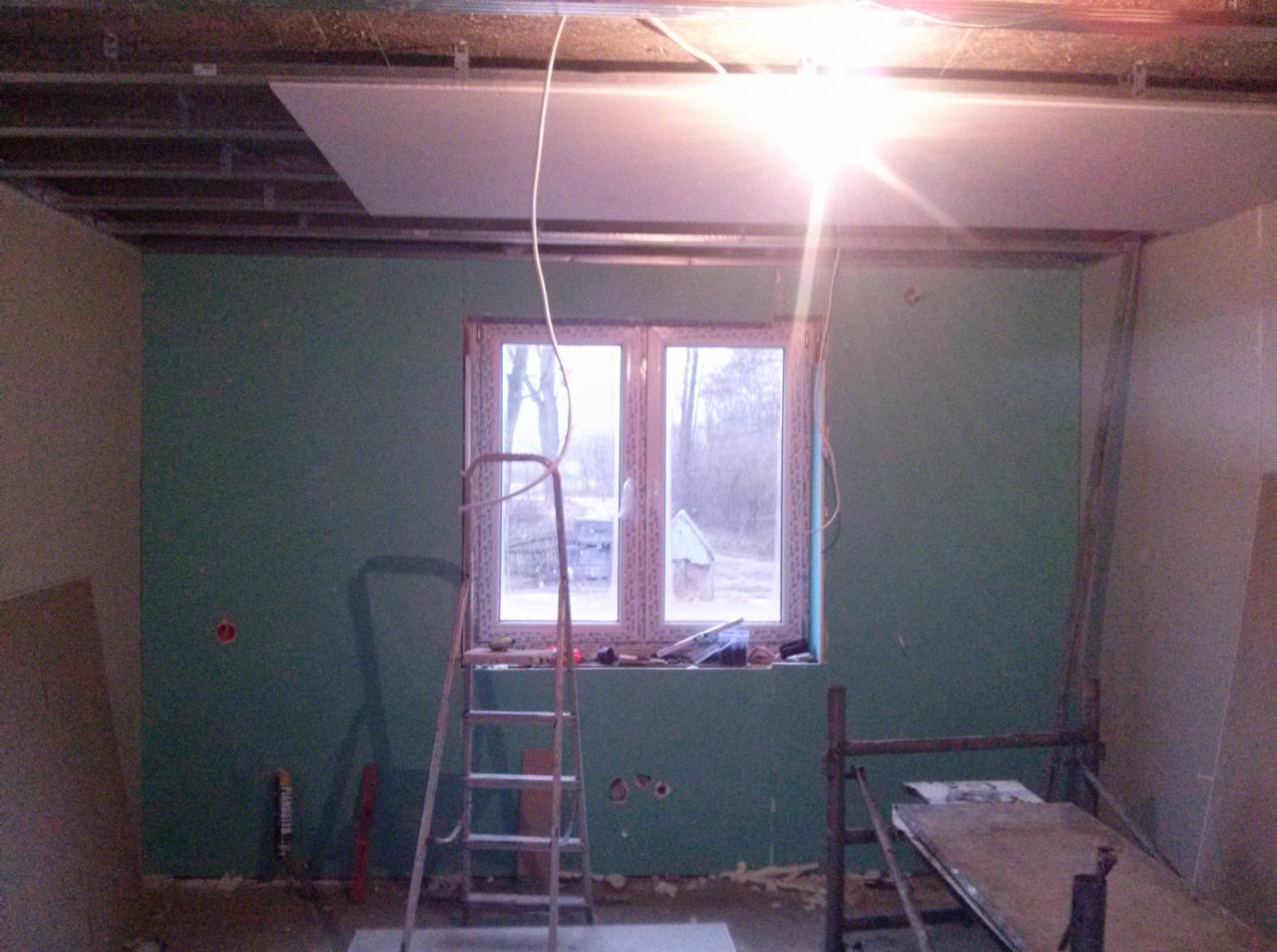 Wnętrze w trakcie montażu płyt karton-gips na suficie i ścianach, widoczne profile, drabina i okno. Prace wykończeniowe w pomieszczeniu z zielonymi ścianami.