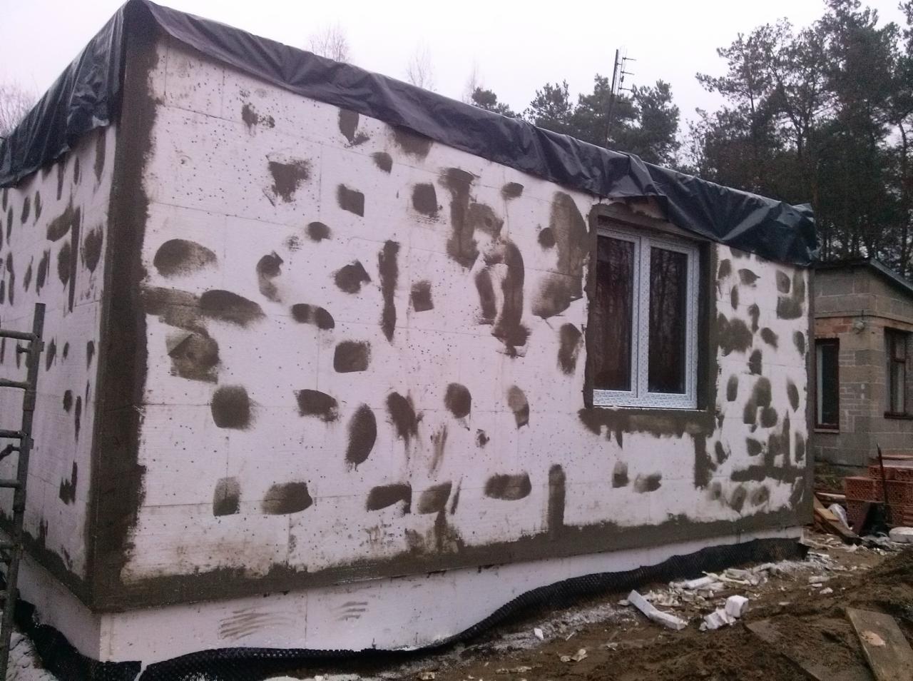 Budynek w trakcie ocieplania styropianem, z widocznymi plackami kleju, zabezpieczony czarną folią na dachu. Nowe okno PCV. W tle drugi budynek.