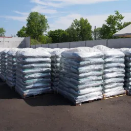 Rzędy worków z opałem na paletach, zabezpieczone folią, na składzie z betonowymi ścianami i zielenią w tle, w jasny słoneczny dzień.