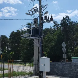 Słup energetyczny z transformatorem i skrzynką przyłączeniową na kostce brukowej, w tle drzewa i błękitne niebo z chmurami.