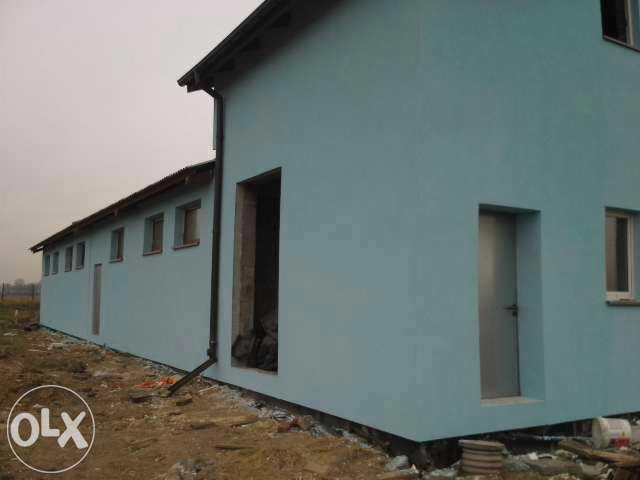 Budynek po remoncie elewacji w odcieniach błękitu, z widocznymi oknami i drzwiami. Nierówny teren wokół budynku. Widoczne elementy wykończeniowe.