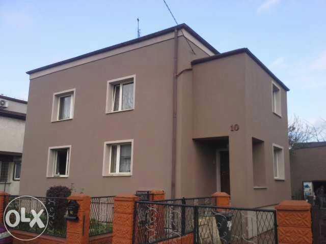 Dwupiętrowy dom z beżową elewacją, widoczny zza ogrodzenia. Na ścianie numer '10'. Okna z białymi ramami. Niebieskie niebo w tle. Budynek po renowacji.