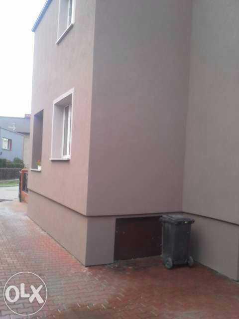 Fragment elewacji domu po remoncie, widoczne okna i cokół. Ciemny śmietnik przy ścianie. Kostka brukowa na podjeździe.