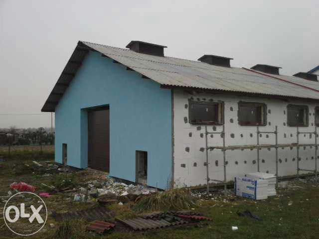 Budynek w trakcie ocieplania: część elewacji pomalowana na niebiesko, druga w trakcie ocieplania z widocznym styropianem i rusztowaniem. Dach z blachy falistej.