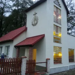 Jasna elewacja budynku z czerwonym dachem, widoczne okna z luksferów, krzyż na fasadzie i fragment drewnianego płotu na pierwszym planie.