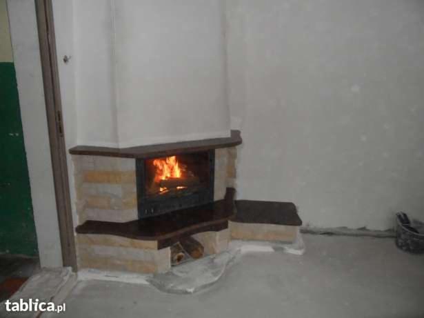 Murowany kominek z palącym się ogniem, obudowany kamieniem i drewnem, stoi przy białej ścianie. Widoczne palenisko i fragment podłogi.