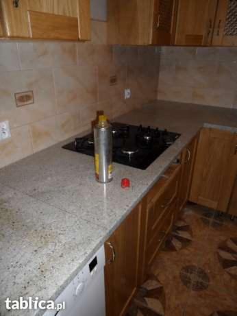 Jasny blat kamienny z kuchenką gazową i szafkami z drewna. Na blacie stoi wysoki spray, a w tle widać beżowe płytki na ścianie i fragment podłogi.