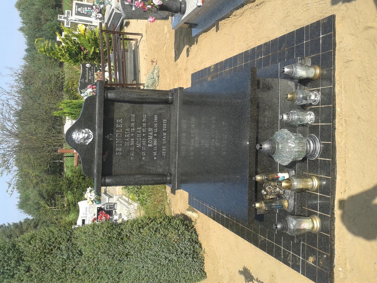 Czarny, polerowany nagrobek z inskrypcjami na cmentarzu, otoczony piaskiem i zielenią, z zapalonymi zniczami na czarnej, kafelkowej podstawie. Ujęcie z boku, w słoneczny dzień.