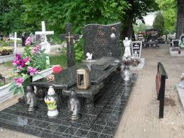 Granitowy nagrobek z krzyżem i lampionem na cmentarzu, otoczony zielenią i innymi nagrobkami. Widok z boku, lekko z góry, prezentujący detale kamieniarskie.