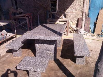 Komplet mebli ogrodowych z szarego kamienia: stół i ławki o surowym, minimalistycznym designie, ustawione na zewnątrz budynku. Widoczne drobne niedoskonałości obróbki kamienia.