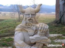 Rzeźba z piaskowca przedstawiająca wikinga z rogatym hełmem, trzymającego miecz. Na drugim planie łąka i budynek z czerwonej cegły.